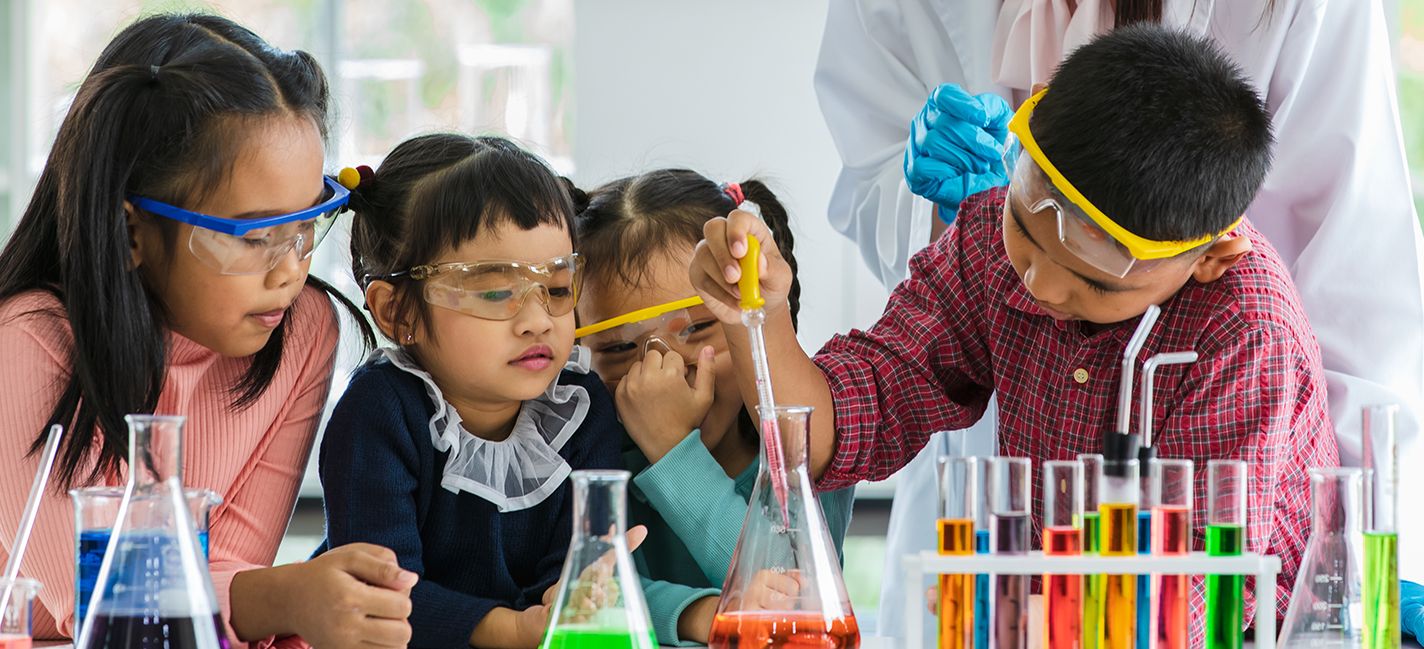 Boy drops something into orange beaker using a pipette in a lab while 3 little girls watch all wearing safety goggles