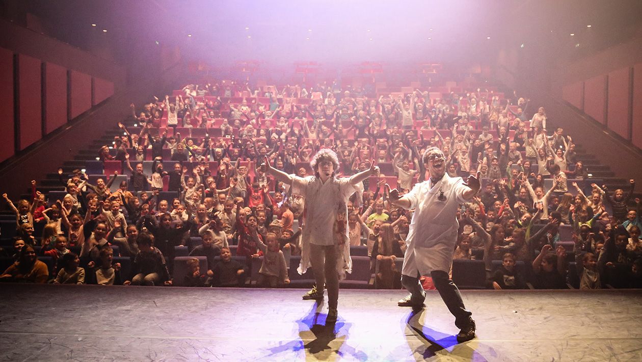 2 Mad scientists in labcoats on stage with their back towards a large excited crowd 